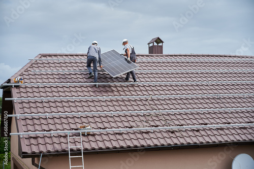 Men technicians carrying photovoltaic solar modul on roof of house. Workmen in helmets installing solar panel system outdoors. Concept of alternative and renewable energy.