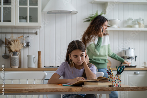 Focused smart teen girl making homework sits at table in dining room doing school extracurricular tasks. Concentrated daughter reading textbooks mother talking on phone standing in kitchen