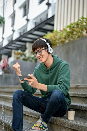 Cheerful Asian male college student wearing headphones and using his smartphone on street stairs © bongkarn