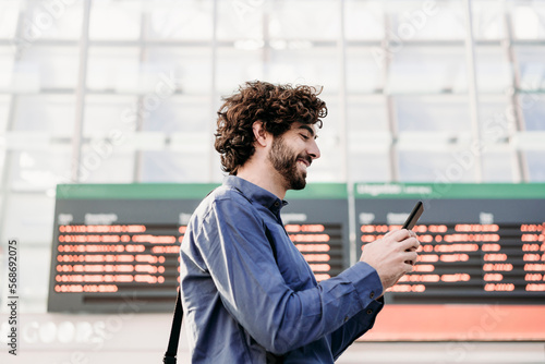 Happy businessman using mobile phone by arrival departure board at station