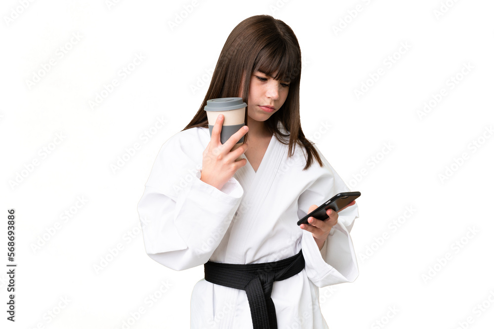 Little Caucasian girl doing karate over isolated background holding coffee to take away and a mobile Little Caucasian girl doing karate over isolated background holding coffee to take away and a mobile