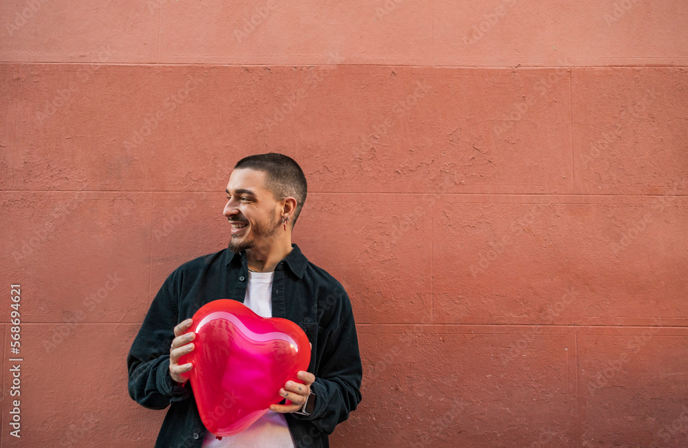 Happy man holding red heart shape balloon in front of wall Happy man holding red heart shape balloon in front of wall