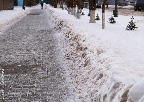 Snowdrifts along a cleared footpath made of paving slabs.A blurry background of a snow-covered city street with cars, trees and people. Without a face