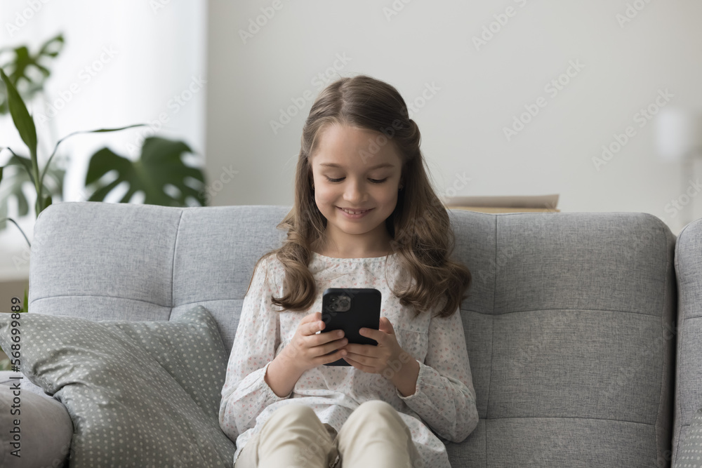 Cheerful little kid girl playing video game on mobile phone, resting on sofa at home, making call to parents, using learning app for education, online communication Cheerful little kid girl playing video game on mobile phone, resting on sofa at home, making call to parents, using learning app for education, online communication