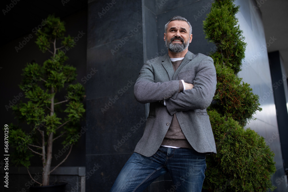a mature satisfied male businessman is standing in a jacket at the entrance to the business center with his arms crossed on his chest a mature satisfied male businessman is standing in a jacket at the entrance to the business center with his arms crossed on his chest