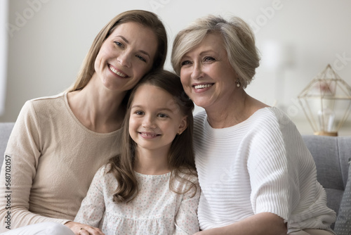 Cheerful cute child, mother and granny hugging on home couch with love, care, tenderness, enjoying closeness, family meeting, warm relationship, leisure together, laughing, smiling, looking away