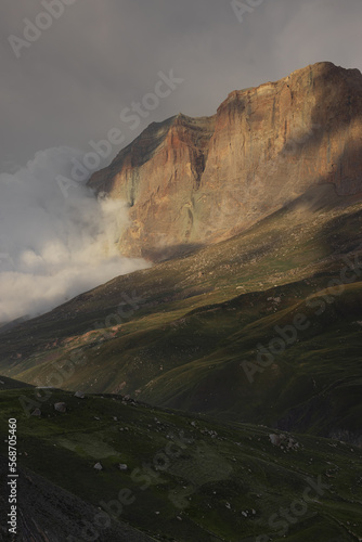 Breathtaking summer mountain landscape - high orange rocky cliff of canyon in in golden sunbeam and shadows flowing fluffy cloud on green velvety hilly slope, grey stormy sky. Majestic view, vertical.