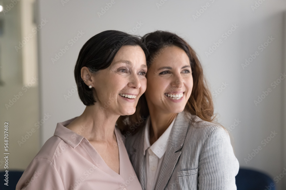 Happy beautiful senior mom and successful adult daughter standing close, hugging with face, cheek touches, looking away with dreamy toothy smiles, laughing. Family portrait Happy beautiful senior mom and successful adult daughter standing close, hugging with face, cheek touches, looking away with dreamy toothy smiles, laughing. Family portrait