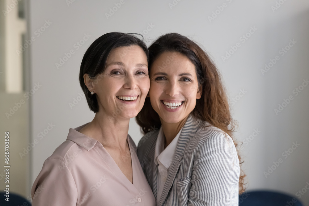 Cheerful senior middle aged mom and pretty brunette daughter woman posing cheek to cheek together, standing close with face touches, looking at camera, smiling, laughing, hugging Cheerful senior middle aged mom and pretty brunette daughter woman posing cheek to cheek together, standing close with face touches, looking at camera, smiling, laughing, hugging