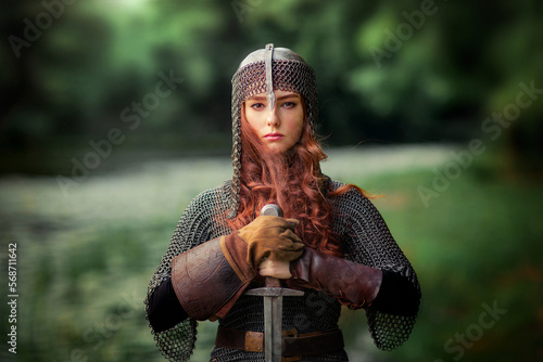 Fototapet Beautiful red haired girl in metal medieval armor dress with sword standing in warlike pose and looking at camera with fearless
