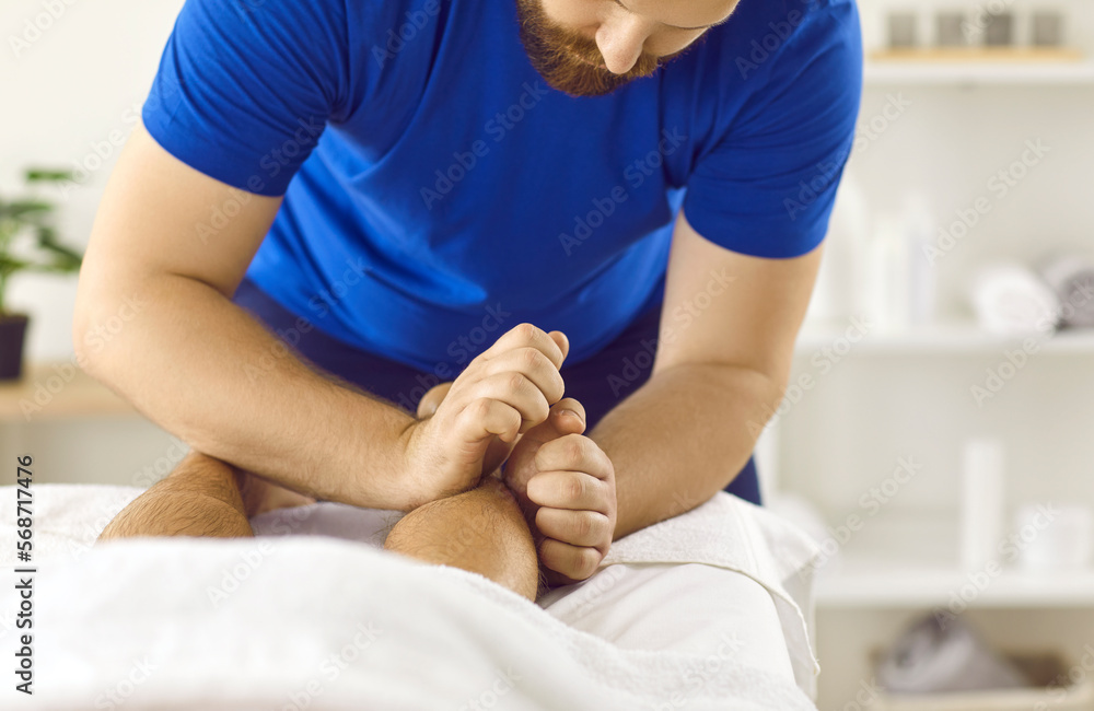 Close up of male therapist massage patient legs help relieve muscle strain after trauma or injury. Man masseur or physiotherapist work with client lying on table in studio or spa. Rehabilitation.