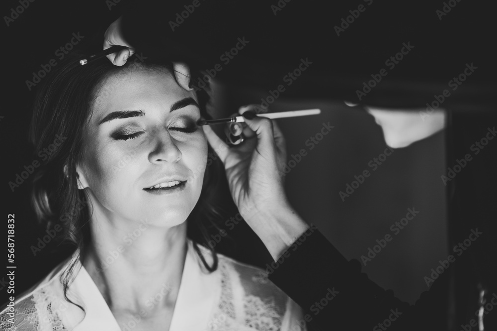 Obraz premium Make-up artist applying eyeshadow on model's eye and holding a brush on background, close up. Young beautiful woman. Black and white photo.