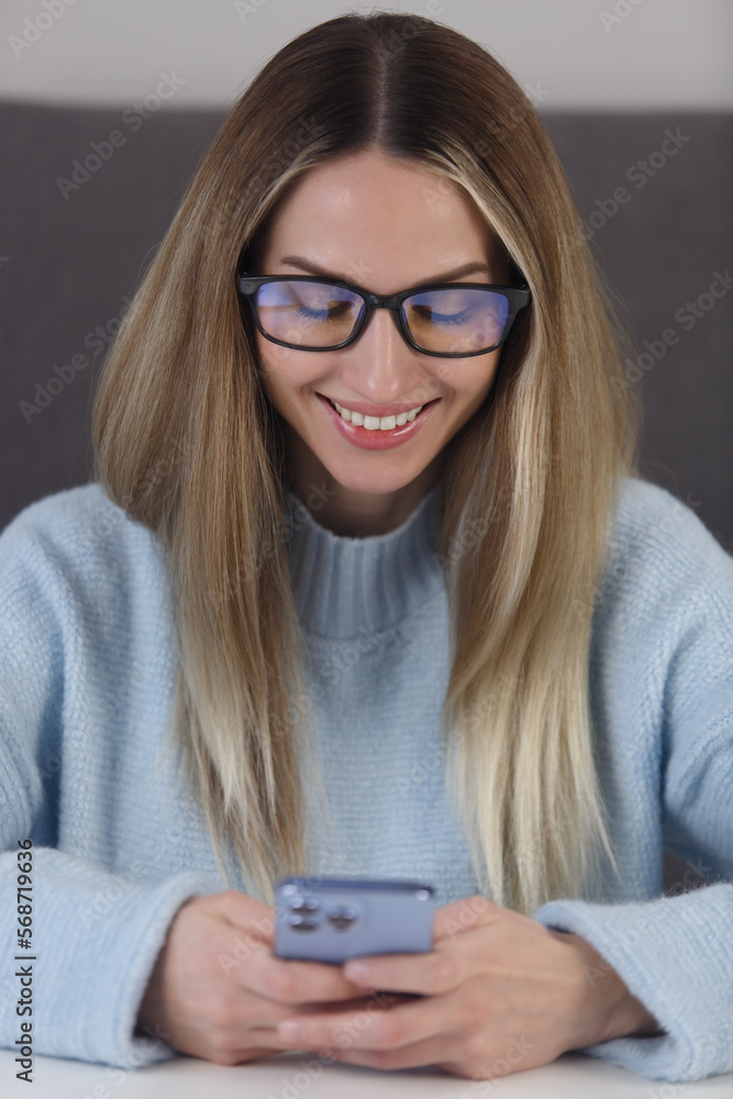 Happy blonde woman in glasses typing a message in modern smart phone app. Closeup portrait of young adult female using mobile phone with a smile