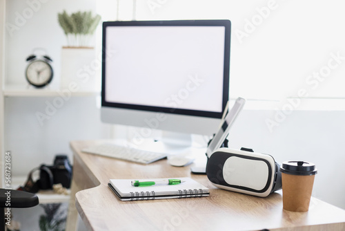 Office desk with modern technology devices. VR headset laying and blank white screen of computer. Mock up and copy space for business at digital era. 