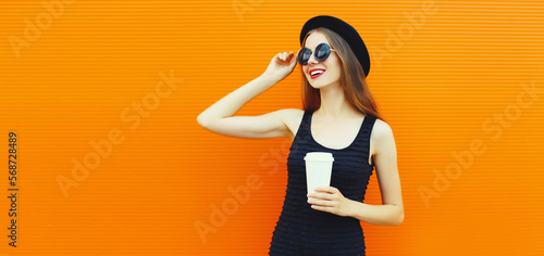 Portrait of happy smiling young woman drinking fresh juice or coffee wearing round black hat on orange background