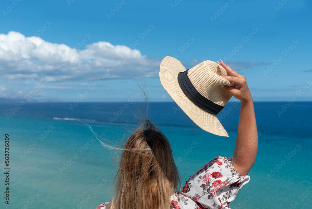 Φωτογραφία Mujer rubia con un vestido de flores y un sombrero blanco de espaldas al cielo azul y al mar turquesa en la costa de la isla turística de Fuerteventura en las Islas Canarias