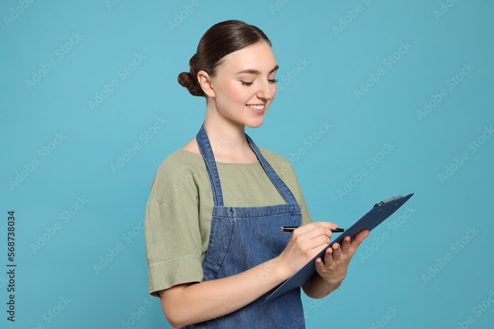 Beautiful young woman in clean denim apron with clipboard on light blue background Beautiful young woman in clean denim apron with clipboard on light blue background