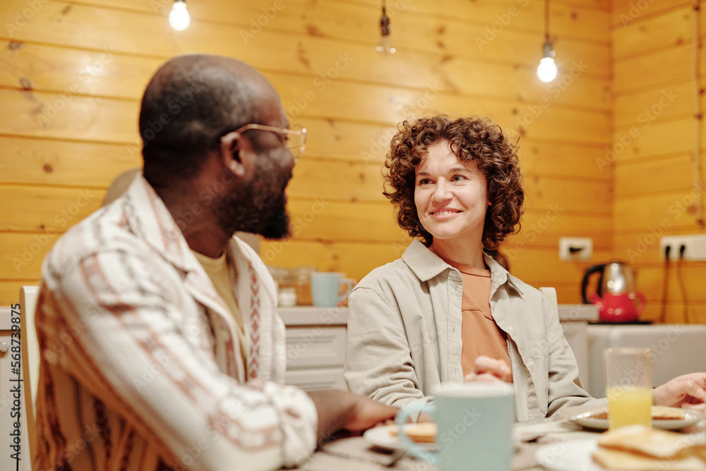 Young smiling brunette woman looking at her African American husband while sitting in front of him by served table during breakfast Young smiling brunette woman looking at her African American husband while sitting in front of him by served table during breakfast