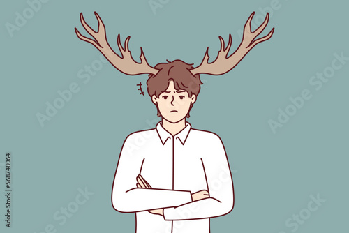 Disgruntled guy with deer horns on head stands with arms crossed and looks at screen with displeasure. Metaphor of man who suffered from infidelity of girlfriend or adultery wife