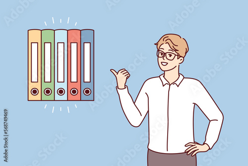 Man office worker points finger at multicolored folders for documents. Guy in business clothes gives advice on finding reports or recommends system for storing financial papers