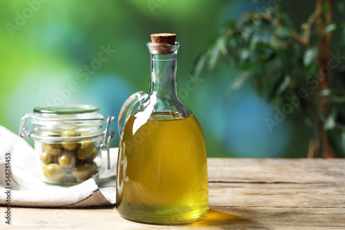 Jug of cooking oil and jar with olives on wooden table against blurred background. Space for text