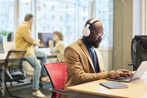 Serious African American employee with headphones sitting by desk in front of laptop and entering new data for business project