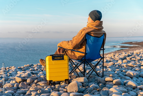 Bearded Man in brown jacket with yellow suitcase relaxing alone and sitting on beach chairs on the seaside at sunrise. Travel Lifestyle concept © Iryna