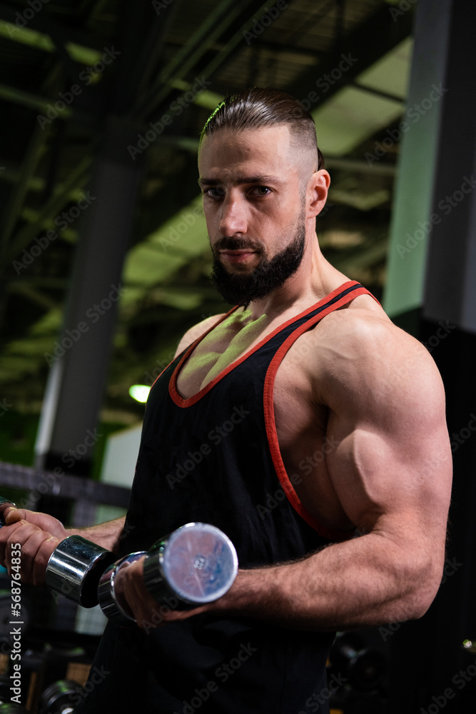 A handsome and brutal athlete with a beard holds dumbbells in his hands and does sports in the gym A handsome and brutal athlete with a beard holds dumbbells in his hands and does sports in the gym