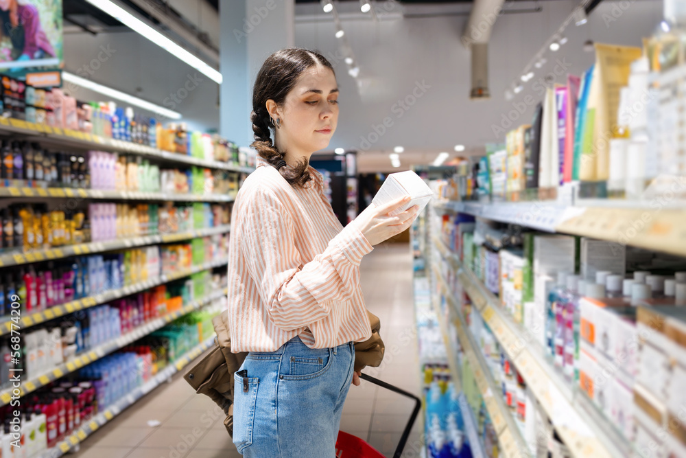 Side view of Caucasian woman choosing cosmetic products in grocery store. Shelves in background. Concept of shopping and consumerism