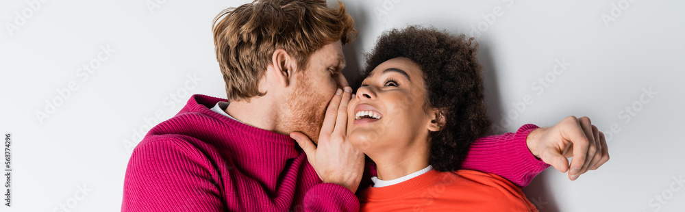top view of redhead man whispering in ear of cheerful african american woman lying on white, banner. top view of redhead man whispering in ear of cheerful african american woman lying on white, banner.