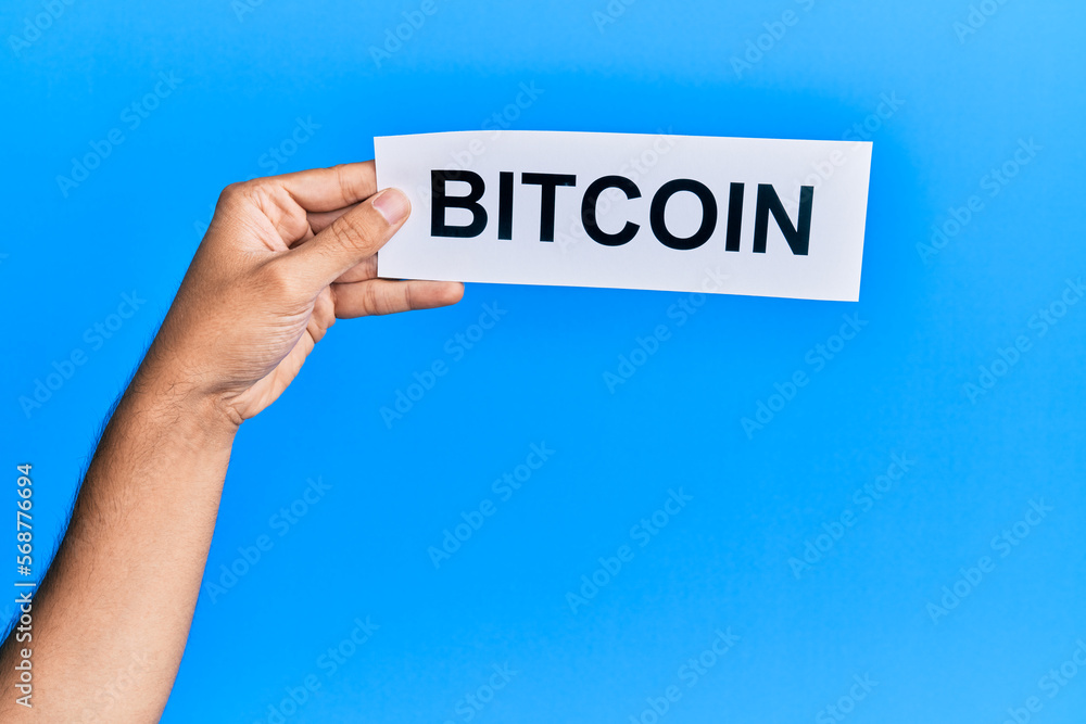 Hand of caucasian man holding paper with bitcoin word over isolated blue background Hand of caucasian man holding paper with bitcoin word over isolated blue background