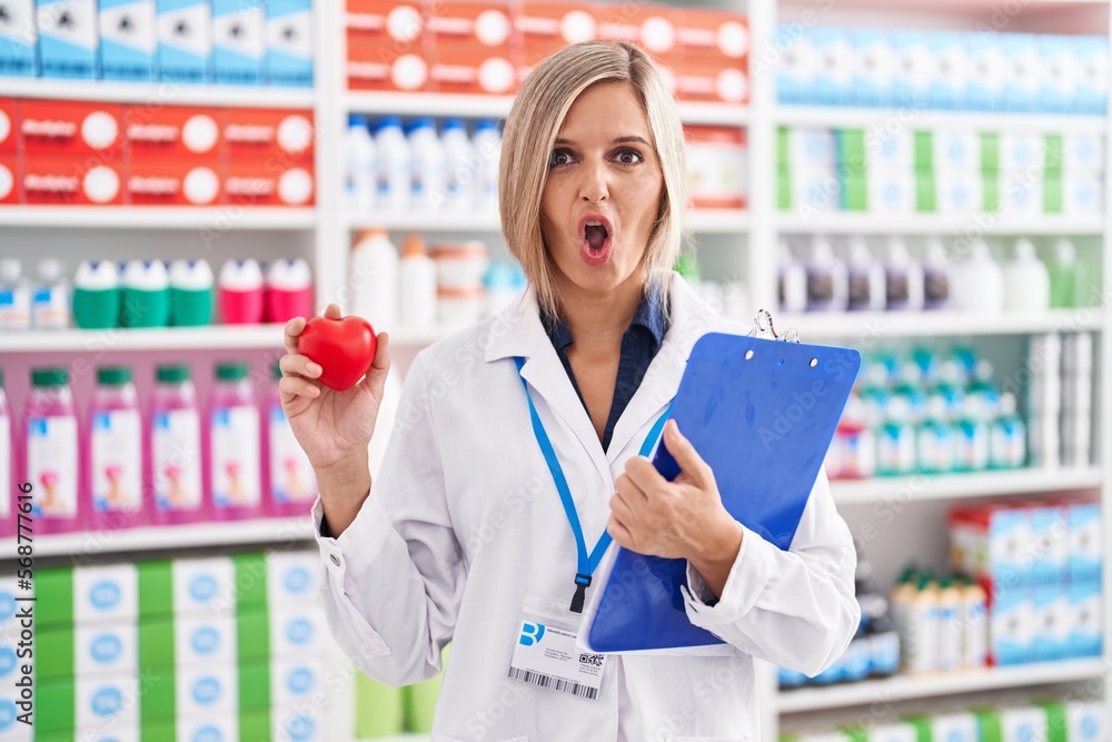 Young woman working at pharmacy drugstore holding heart afraid and shocked with surprise and amazed expression, fear and excited face.