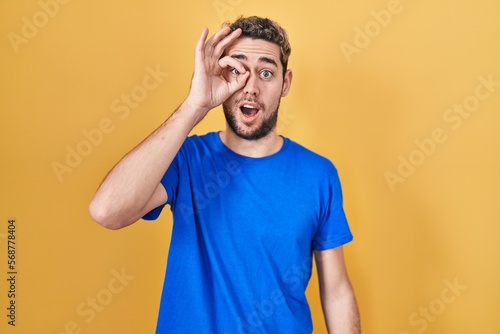 Hispanic man with beard standing over yellow background doing ok gesture shocked with surprised face, eye looking through fingers. unbelieving expression.