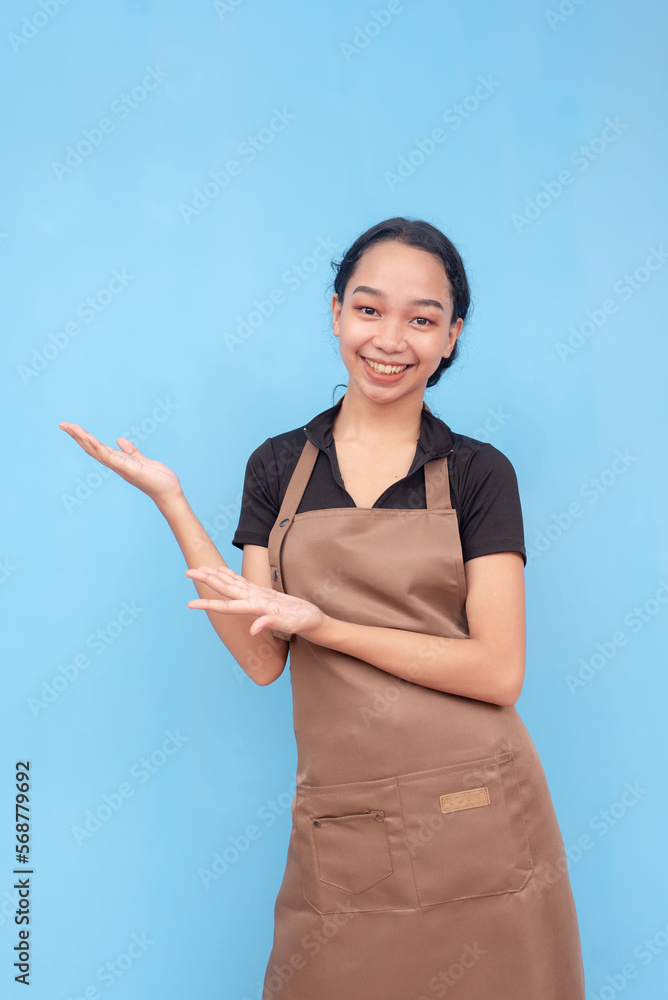 A young female barista of asian descent wearing a brown apron and black shirt presenting something to the left. Against a light blue background. A young female barista of asian descent wearing a brown apron and black shirt presenting something to the left. Against a light blue background.