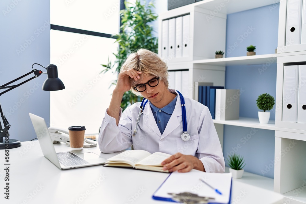 Young blond man doctor using laptop reading book at clinic Young blond man doctor using laptop reading book at clinic