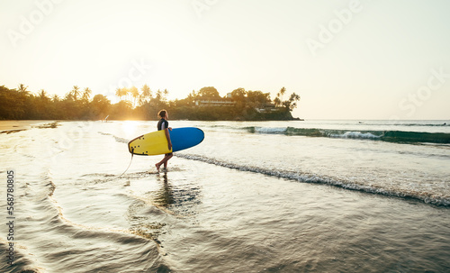 Teen boy with blue and yellow surfboard entering the waves for surfing with sunset rays on palm trees grove. Happy childhood and active sporty people vacation time concept. photo