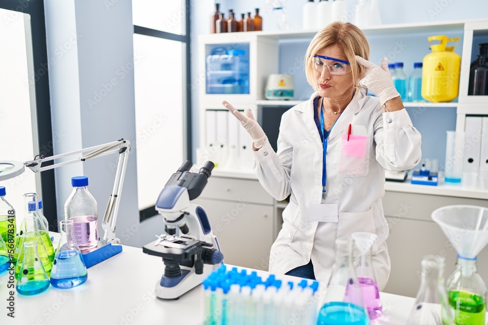 Middle age blonde woman working at scientist laboratory confused and annoyed with open palm showing copy space and pointing finger to forehead. think about it. Middle age blonde woman working at scientist laboratory confused and annoyed with open palm showing copy space and pointing finger to forehead. think about it.