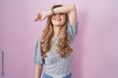 Beautiful blonde woman standing over pink background covering eyes with arm smiling cheerful and funny. blind concept.