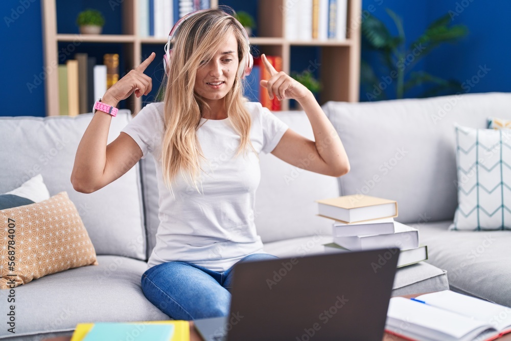 Young blonde woman studying using computer laptop at home smiling pointing to head with both hands finger, great idea or thought, good memory Young blonde woman studying using computer laptop at home smiling pointing to head with both hands finger, great idea or thought, good memory