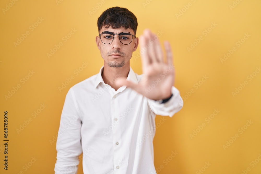 Young hispanic man standing over yellow background doing stop sing with palm of the hand. warning expression with negative and serious gesture on the face. Young hispanic man standing over yellow background doing stop sing with palm of the hand. warning expression with negative and serious gesture on the face.
