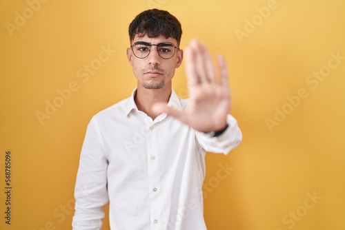 Young hispanic man standing over yellow background doing stop sing with palm of the hand. warning expression with negative and serious gesture on the face.