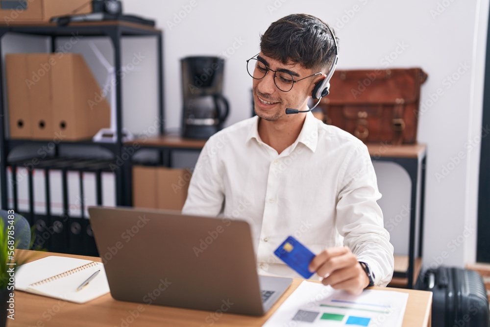 Young hispanic man working using computer laptop holding credit card looking away to side with smile on face, natural expression. laughing confident. Young hispanic man working using computer laptop holding credit card looking away to side with smile on face, natural expression. laughing confident.