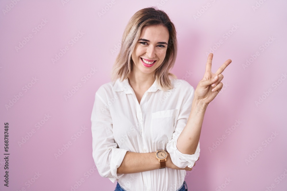 Young beautiful woman standing over pink background smiling with happy face winking at the camera doing victory sign with fingers. number two. Young beautiful woman standing over pink background smiling with happy face winking at the camera doing victory sign with fingers. number two.