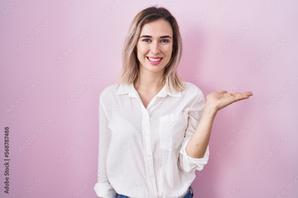 Young beautiful woman standing over pink background smiling cheerful presenting and pointing with palm of hand looking at the camera. Young beautiful woman standing over pink background smiling cheerful presenting and pointing with palm of hand looking at the camera.