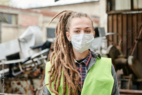 Portrait of volunteer in medical mask and neon vest standing at scrapyard and looking at camera