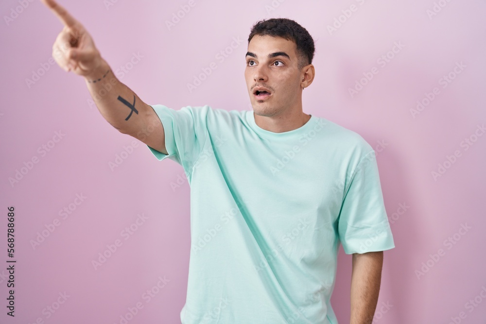 Handsome hispanic man standing over pink background pointing with finger surprised ahead, open mouth amazed expression, something on the front Handsome hispanic man standing over pink background pointing with finger surprised ahead, open mouth amazed expression, something on the front