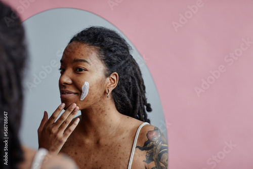 Young black woman with acne scars using face cream looking in mirror photo