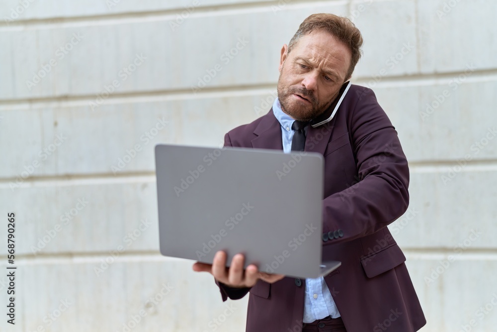 Middle age man business worker talking on smartphone using laptop at street Middle age man business worker talking on smartphone using laptop at street