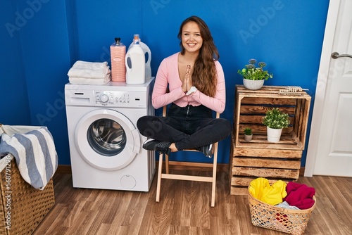 Young beautiful hispanic woman doing yoga exercise waiting for washing machine at laundry room