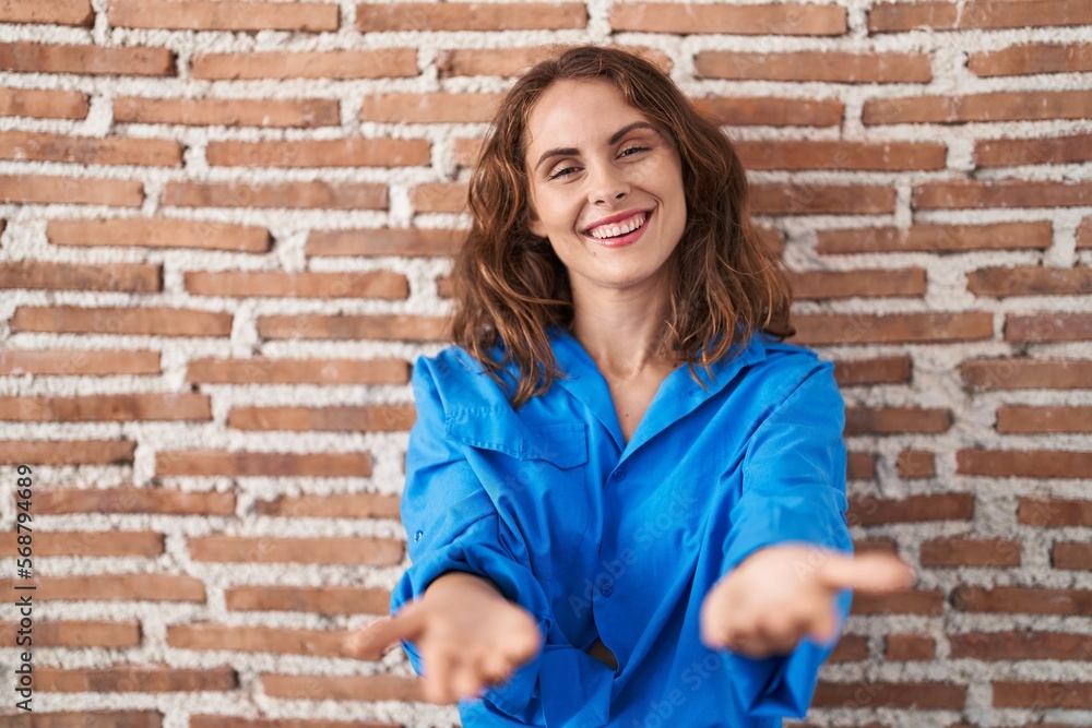 Beautiful brunette woman standing over bricks wall smiling cheerful offering hands giving assistance and acceptance. Beautiful brunette woman standing over bricks wall smiling cheerful offering hands giving assistance and acceptance.